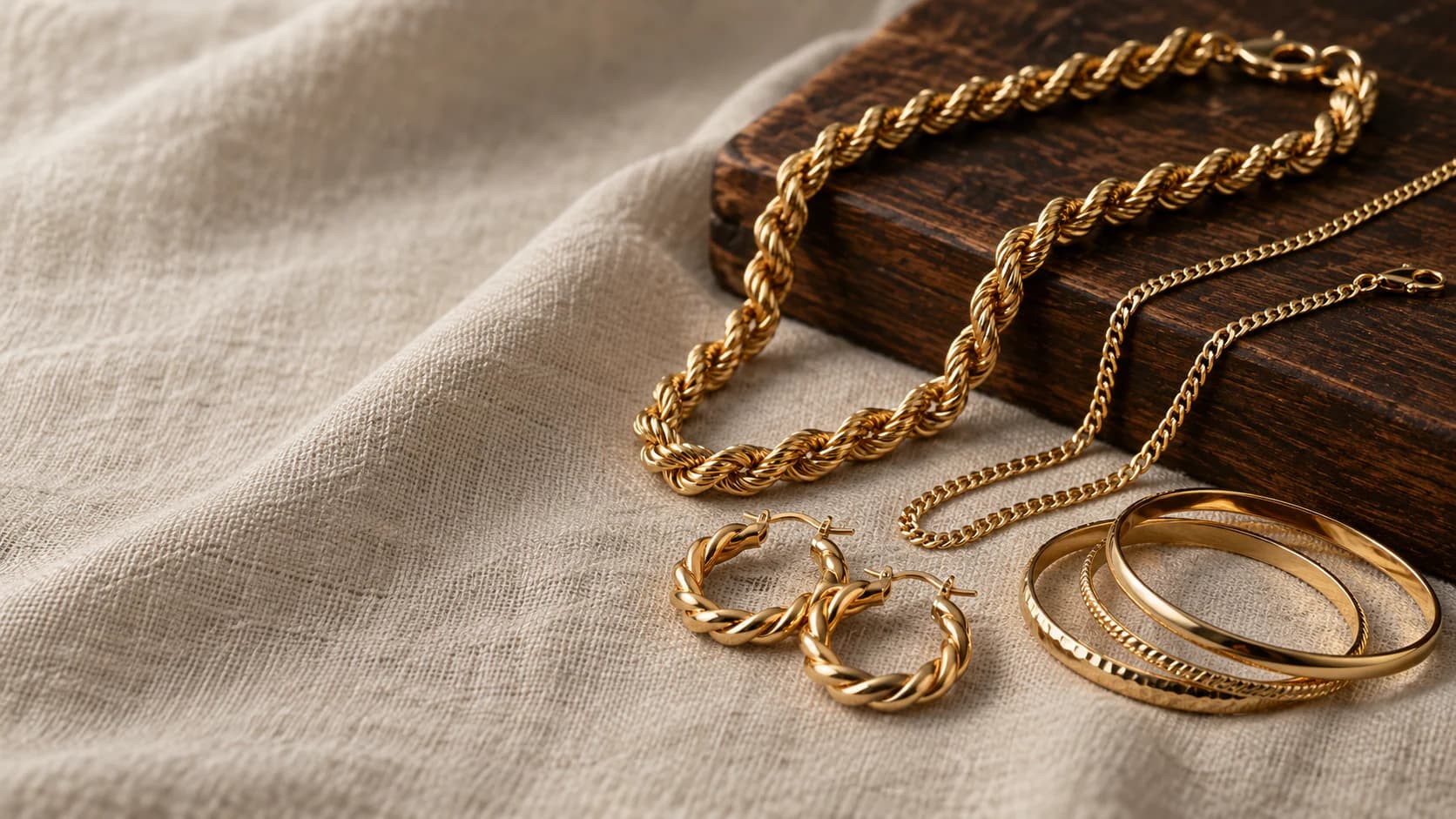 Layered 18ct gold chains and a twisted hoop earring on a dark velvet tray under warm directional light